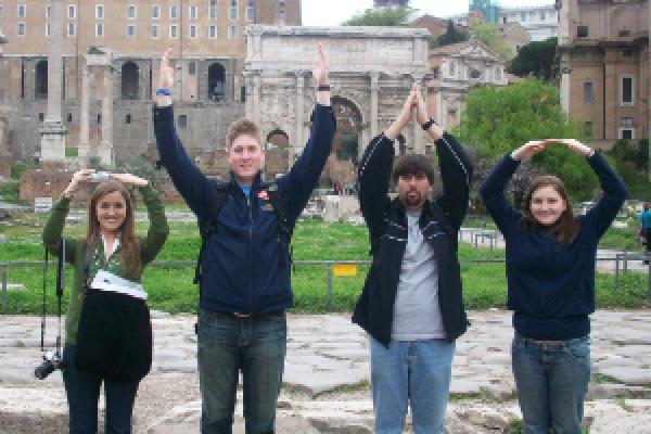 Students make an Ohio State symbol with their arms in front of Roman ruins.