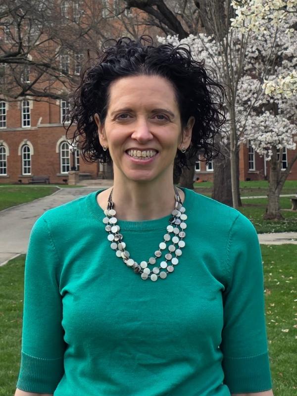 Photo of Megan Nutzman, wearing a green sweater with a white and silver necklace. She is standing outside with a tree and a building in the background. She has dark, curly hair.